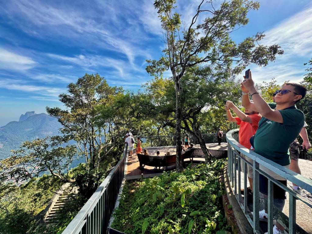 Homens tiram fotos com celulares em um mirante arborizado, com montanhas e céu azul com nuvens ao fundo