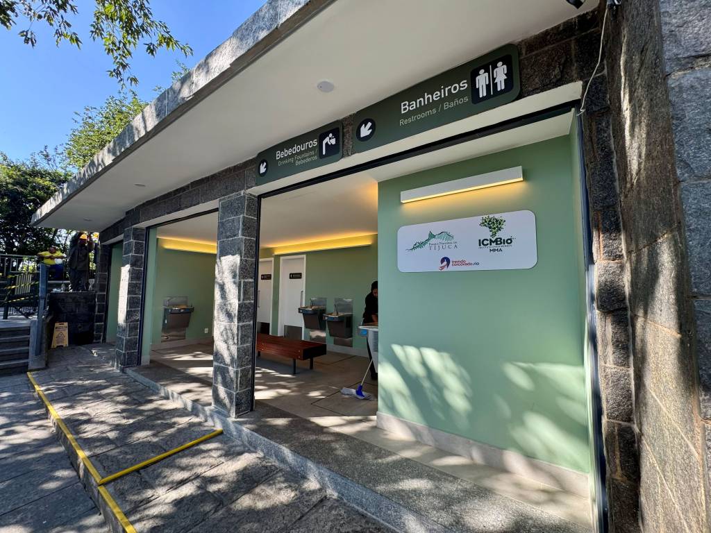 Entrada de um prédio de granito com paredes verde-claro, indicando Bebedouros e Banheiros em placas escuras. Há uma rampa de acesso com faixa amarela, bebedouros de aço e um banco de madeira no interior. Placas com logos Tijuca e ICMBio estão na parede. Um funcionário de colete amarelo aparece ao fundo