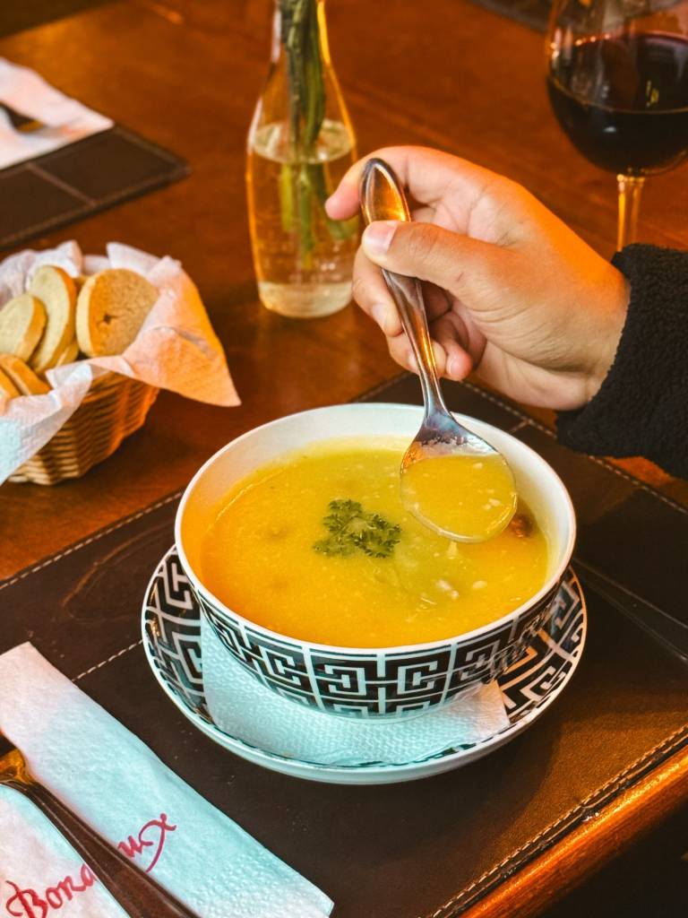Mão de uma pessoa segurando uma colher com sopa amarela, sobre uma tigela preta e branca com a mesma sopa, decorada com salsa. Ao fundo, pães em uma cesta, uma garrafa com planta e uma taça de vinho tinto