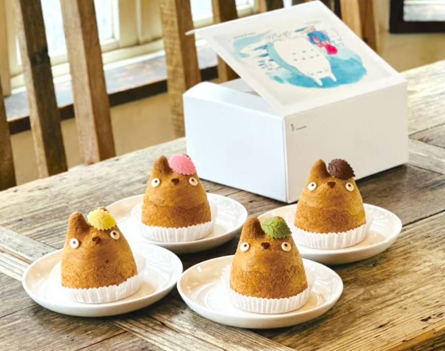 Quatro doces em formato de Totoro, com orelhas e olhos, decorados com folhas coloridas, sobre pratos brancos em uma mesa de madeira. Ao fundo, uma caixa branca aberta com ilustração azul e rosa