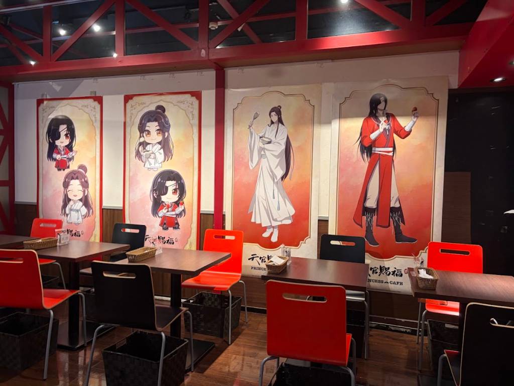 Interior de um café com mesas de madeira e cadeiras vermelhas e pretas. Nas paredes, pôsteres de personagens de anime: dois homens em trajes tradicionais, um branco e outro vermelho, e quatro versões chibi de outros personagens.