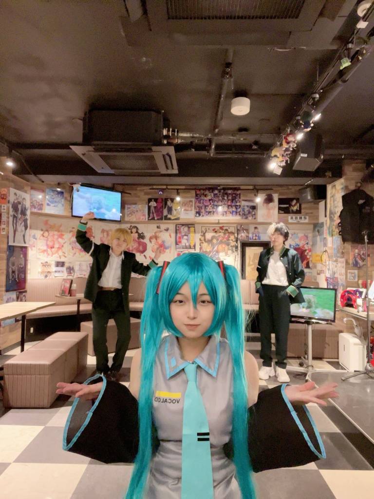 Pessoa fantasiada de Hatsune Miku com peruca azul-turquesa e roupa cinza, gravata azul, braços abertos, olhando para frente. Ao fundo, duas pessoas fantasiadas, uma com cabelo loiro e terno preto, braços erguidos, e outra com cabelo escuro e jaqueta preta, mãos nos bolsos. O ambiente é um café temático com pôsteres de anime nas paredes e televisores exibindo imagens coloridas