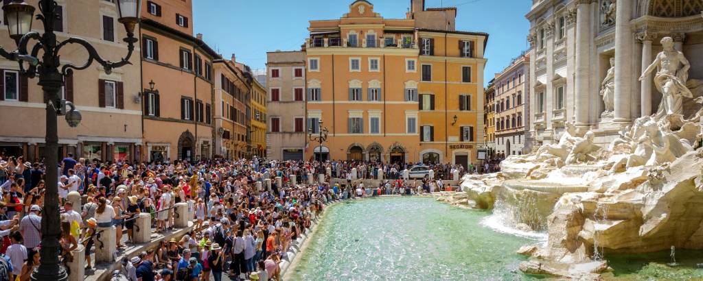 Multidão de turistas em frente à Fontana di Trevi em Roma, com a água azul-esverdeada da fonte e edifícios históricos de tons pastel ao fundo, sob um céu azul claro