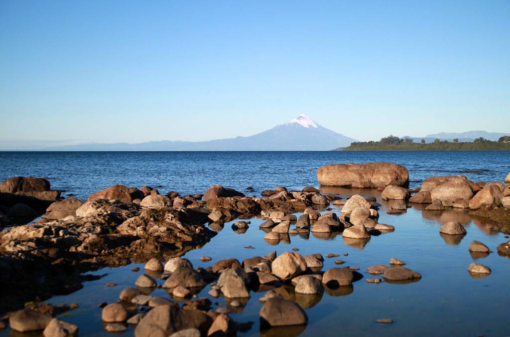 Pedras escuras na margem de um lago azul, refletindo o céu claro. Ao fundo, um vulcão com pico nevado e montanhas distantes sob o horizonte