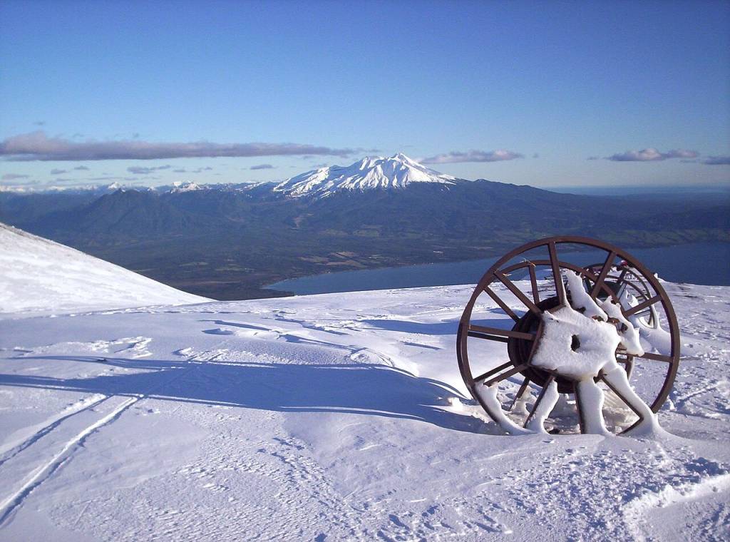 Paisagem montanhosa nevada com um carretel de metal enferrujado coberto de neve em primeiro plano. Ao fundo, um lago azul e montanhas com picos brancos sob um céu claro