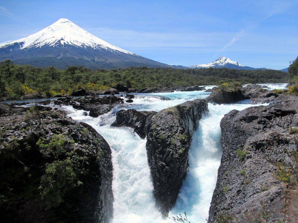 Cachoeiras de Petrohué com águas azul-turquesa fluindo entre rochas escuras, vegetação verde e amarela, e o vulcão Osorno com pico nevado ao fundo sob céu azul