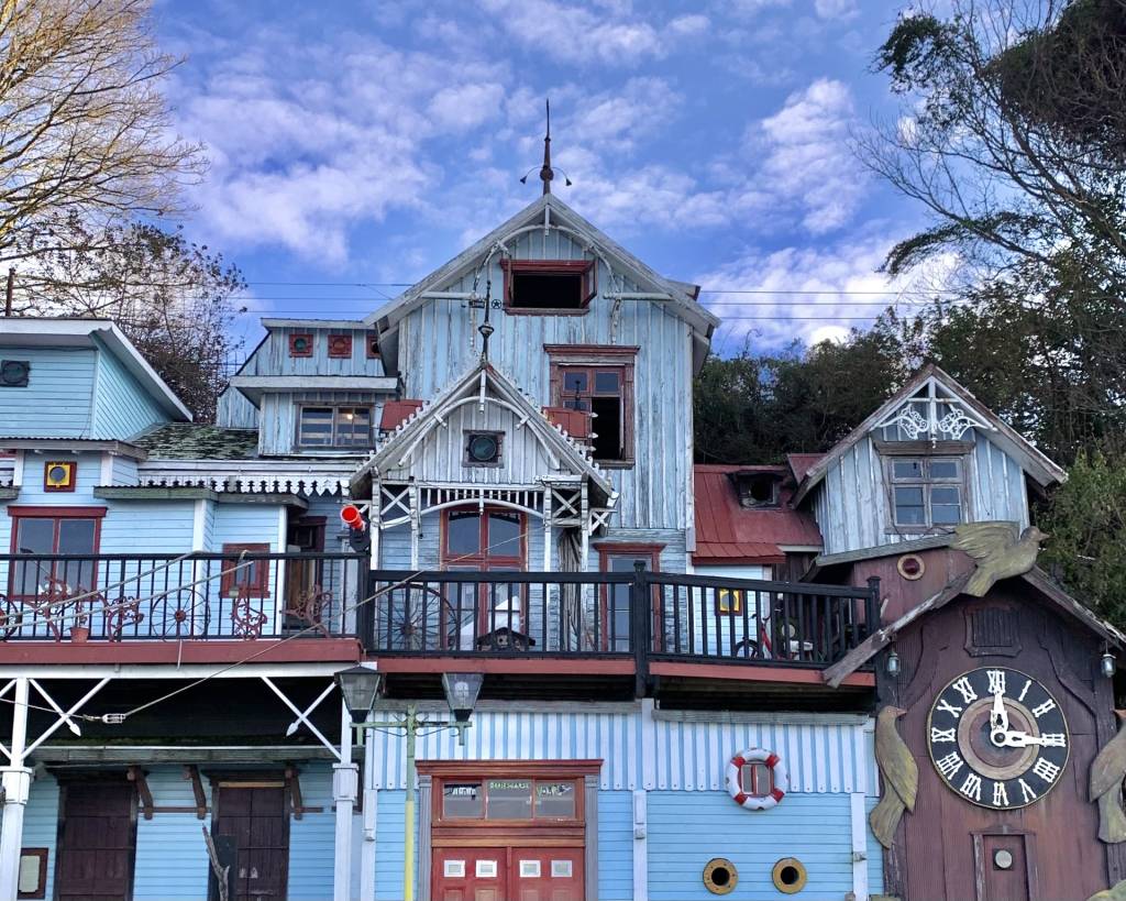 Fachada de uma casa azul-clara com detalhes brancos e vermelhos, telhados de diferentes alturas, varandas com grades pretas e um grande relógio de cuco de madeira escura com números romanos à direita, sob um céu azul com nuvens esparsas. Foto sob licença CC BY-SA 4.0