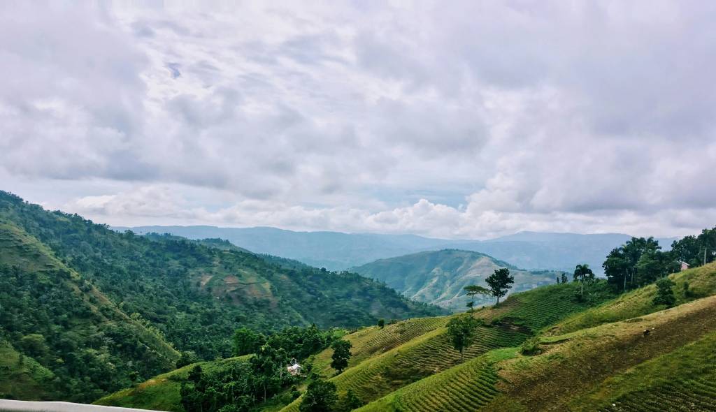 Paisagem montanhosa com colinas verdes e vales profundos, algumas encostas com plantações em terraços. O céu está nublado com nuvens brancas e cinzas, e montanhas distantes aparecem em tons de azul claro. Há árvores esparsas e vegetação densa em algumas áreas