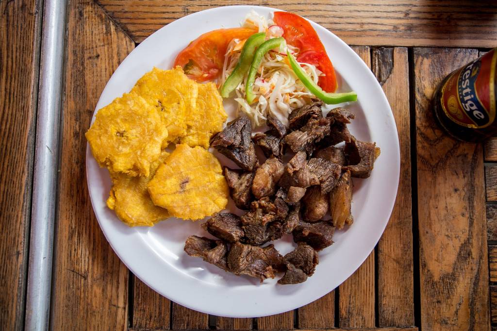 Prato com carne frita em cubos, tostones de banana-da-terra e salada de repolho com tomate e pimentão verde, sobre mesa de madeira rústica, com garrafa de cerveja Prestige ao lado