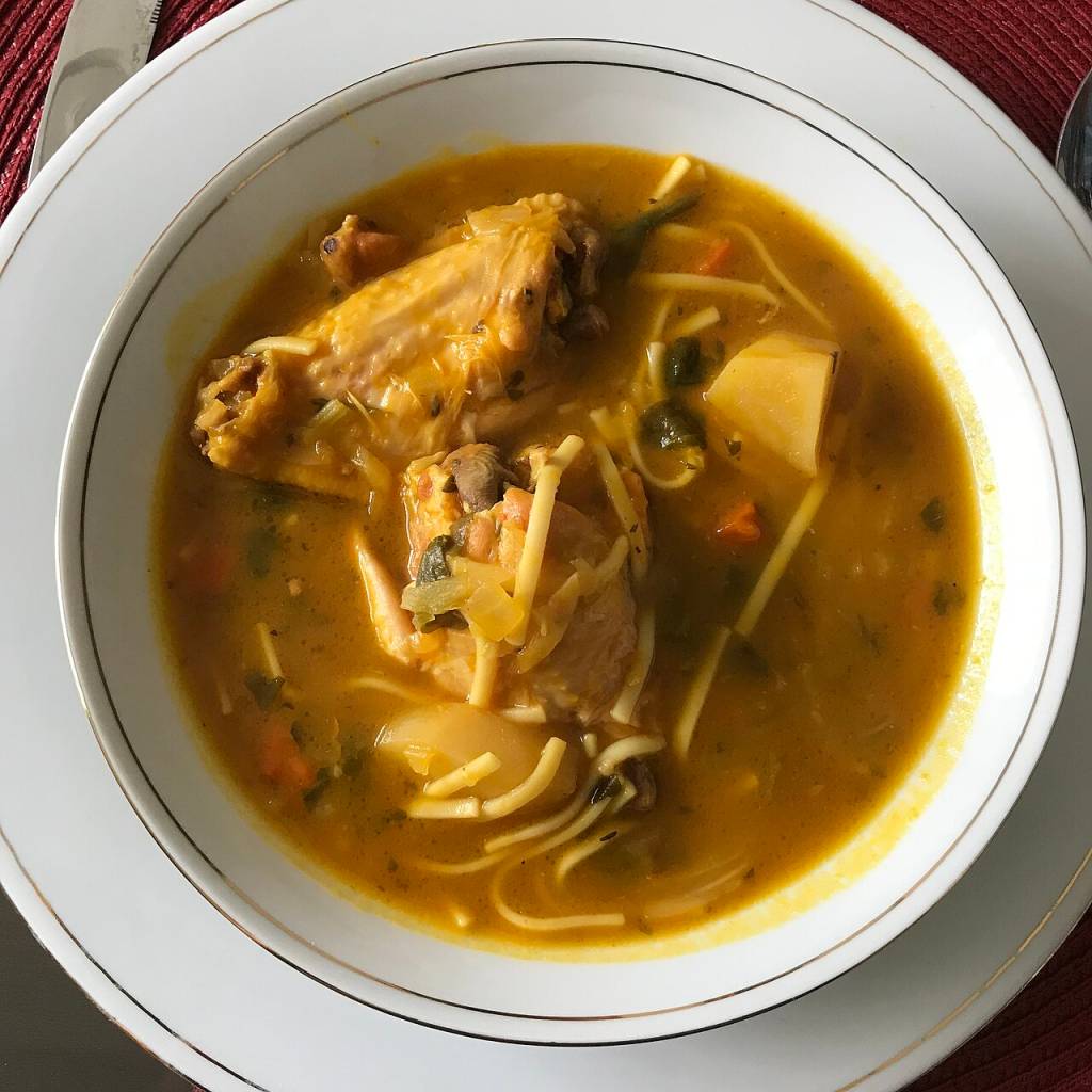 Sopa de galinha com macarrão, batata, cenoura e folhas verdes em caldo amarelo, servida em tigela branca com borda dourada sobre prato branco