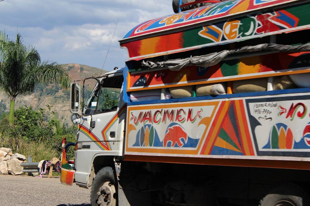 Caminhão branco com carroceria colorida, decorada com padrões geométricos e a palavra JACMEL, parado em uma estrada rural com montanhas e vegetação ao fundo