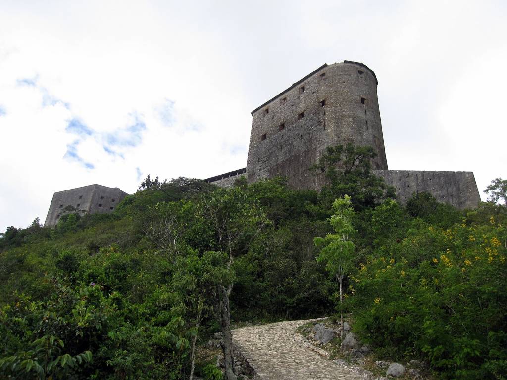 Fortaleza de pedra cinza imponente no topo de uma colina verdejante, com uma estrada de paralelepípedos serpenteando em primeiro plano, sob um céu nublado