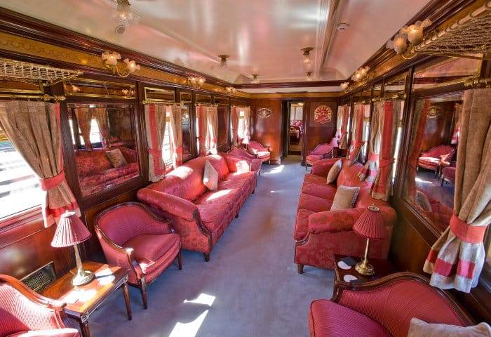 Interior luxuoso de um vagão de trem com sofás e poltronas vermelhas, mesas laterais com abajures, cortinas bege e vermelhas, e detalhes em madeira escura e dourado, criando um ambiente clássico e elegante
