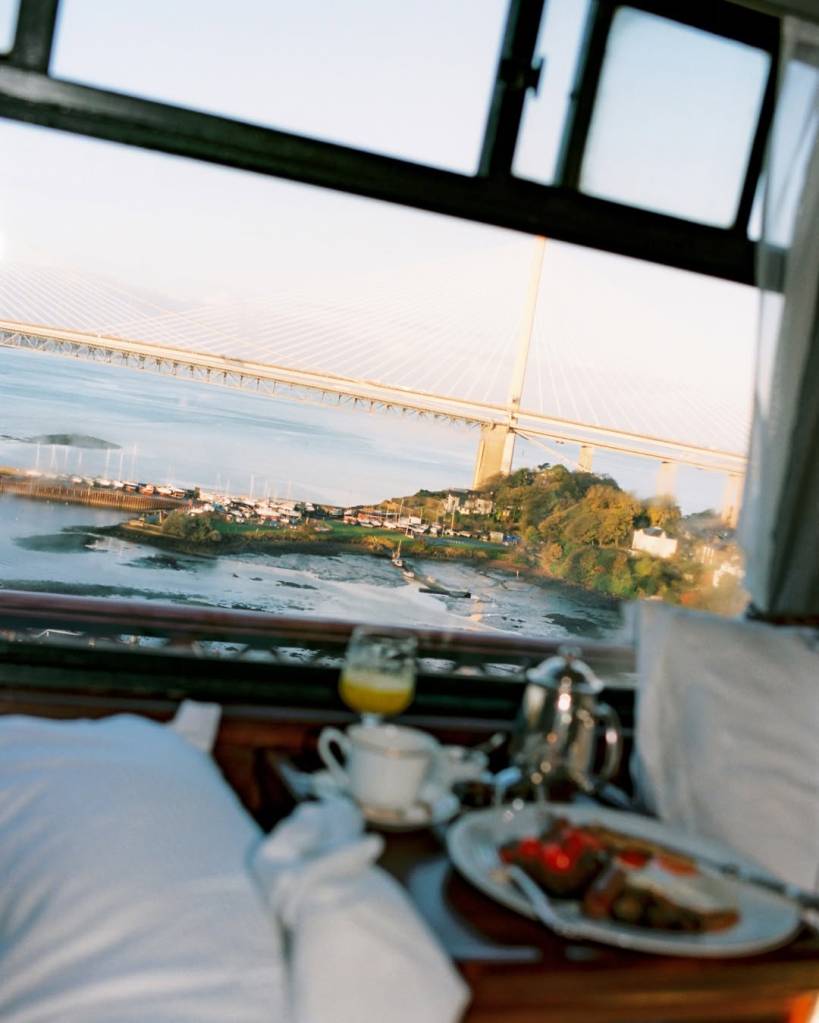 Café da manhã com suco, xícara e bule de prata, servido em uma mesa de madeira escura, com vista para uma ponte estaiada sobre um rio com margens arborizadas e casas, sob céu claro