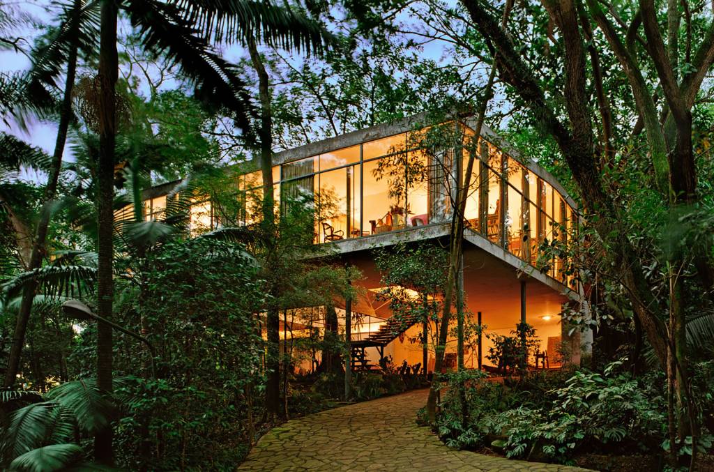 Casa de arquitetura moderna com paredes de vidro, iluminada internamente em tons quentes, cercada por densa vegetação tropical e palmeiras. Um caminho de pedras serpenteia até a entrada, revelando uma escada externa. O céu azul escuro sugere o entardecer.