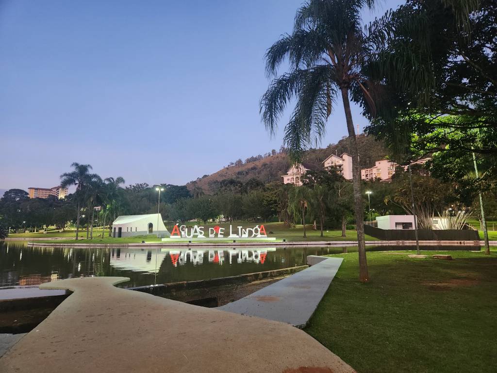 Vista noturna do lago de Águas de Lindoia, com letreiro vermelho ÁGUAS DE LINDOIA refletido na água. Ao fundo, montanhas com prédios e vegetação, e um céu azul escuro. Em primeiro plano, gramado verde e um caminho de concreto.