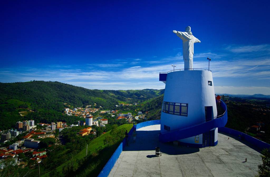 Estátua branca do Cristo Redentor de braços abertos sobre uma base azul-clara, com vista panorâmica de uma cidade em um vale verdejante sob céu azul com nuvens esparsas