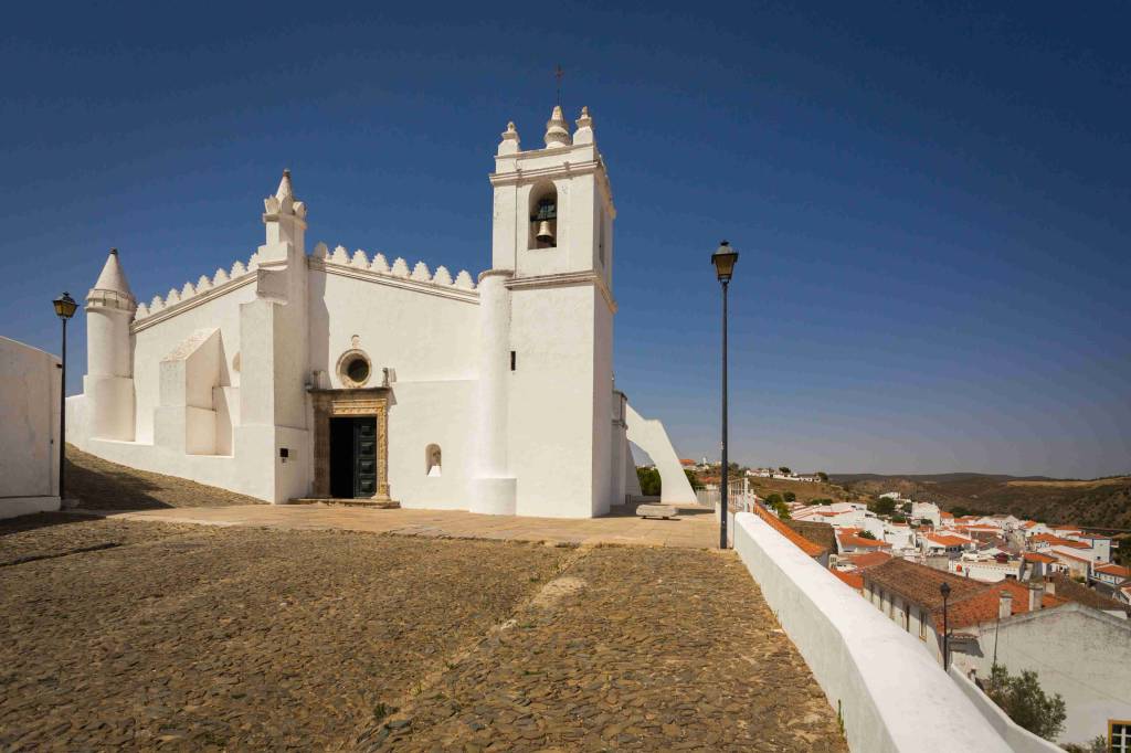 Igreja branca com torre sineira e telhado serrilhado, em praça de paralelepípedos. À direita, um muro branco e casas com telhados de terracota se estendem por uma colina, sob céu azul claro