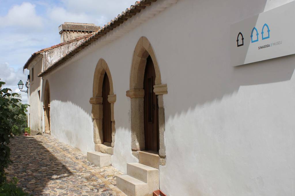 Rua de paralelepípedos com uma parede branca à direita, duas portas de madeira com arcos de pedra e degraus. Um letreiro branco com ícones azuis e pretos e o texto SINAGOGA // MUSEU está na parede. À esquerda, vegetação e um poste de luz antigo. O céu tem nuvens esparsas
