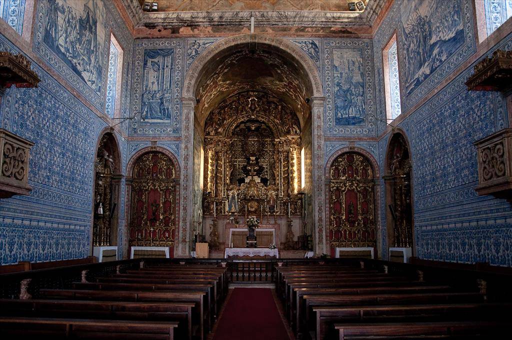 Interior de uma igreja colonial com paredes revestidas por azulejos azuis e brancos, retratando cenas bíblicas. O altar dourado e ricamente ornamentado brilha ao fundo, iluminado por janelas laterais. Bancos de madeira escura preenchem o espaço central, com um tapete vermelho no corredor.