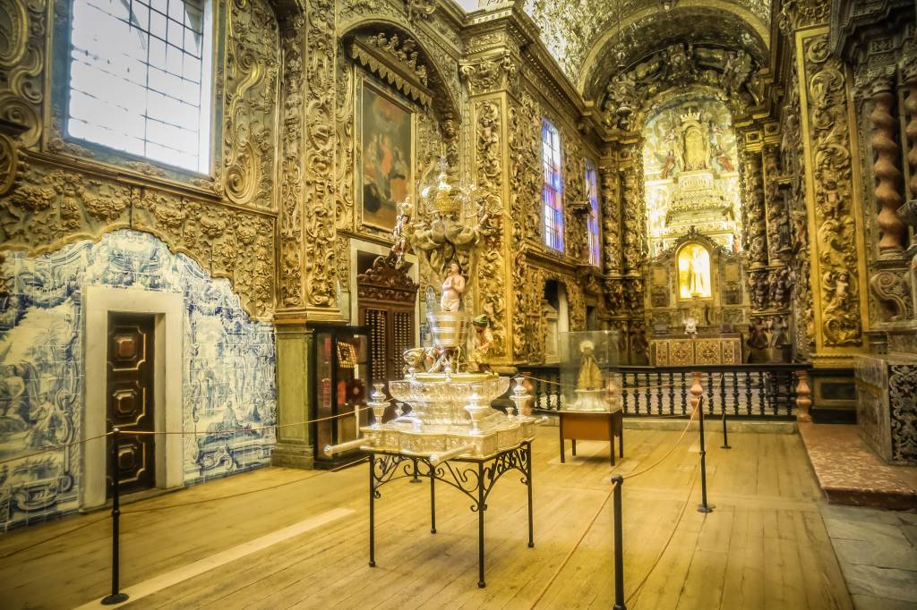 Interior de uma igreja barroca ricamente decorada com talha dourada e azulejos azuis e brancos. O teto e as paredes exibem detalhes ornamentados em ouro, com pinturas e janelas altas. No centro, um altar principal dourado e iluminado. À esquerda, uma parede com azulejos portugueses e uma porta de madeira escura. No primeiro plano, um objeto cerimonial prateado sobre uma mesa de ferro forjado, e uma estátua religiosa em uma vitrine de vidro. O chão é de madeira clara, com cordões de isolamento delimitando o espaço. A atmosfera é de grandiosidade e história
