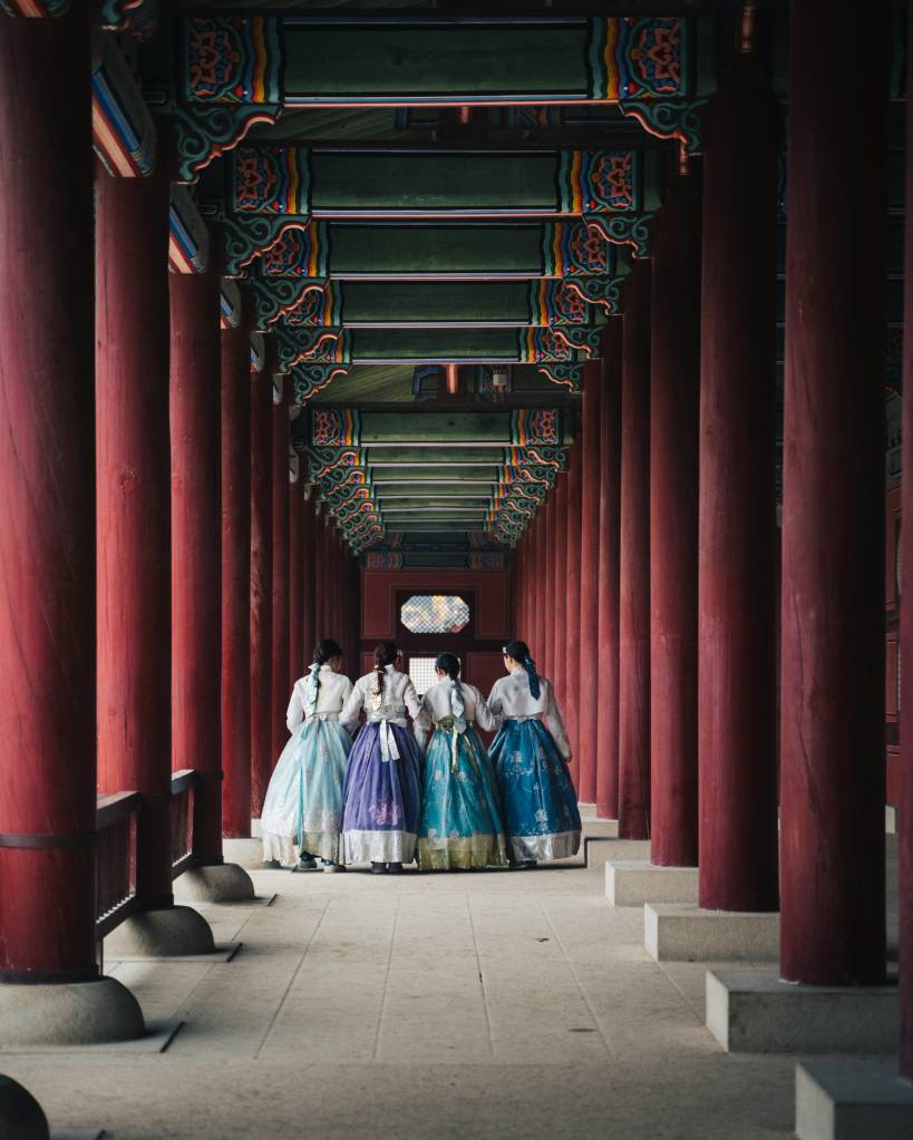 Quatro mulheres de costas, vestindo hanboks coloridos (azul-claro, roxo, turquesa e azul-escuro), caminham por um corredor com pilares vermelhos e teto decorado em tons de verde e laranja, em um palácio coreano