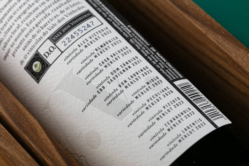 Rótulo de vinho branco com informações sobre a Denominação de Origem Vale dos Vinhedos, número de registro e lista de vinícolas participantes com suas variedades e safras, como Merlot 2022 e Cabernet Sauvignon 2021. Um código de barras aparece na lateral direita, sobre um fundo de madeira escura e um fundo verde na parte superior