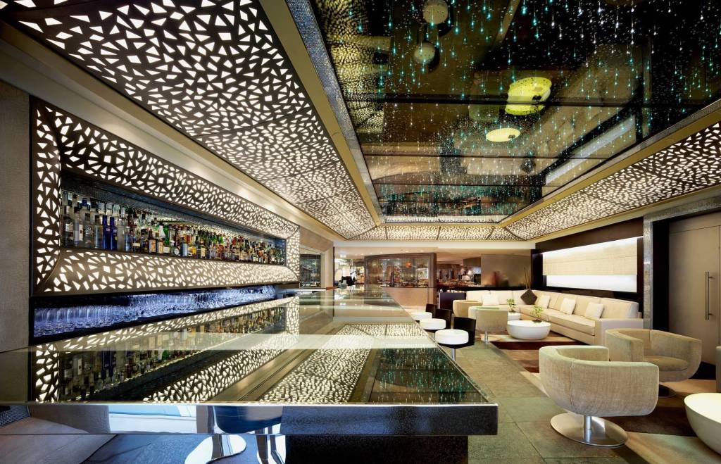 Bar moderno com balcão espelhado, prateleiras de bebidas iluminadas e teto com recortes geométricos e luzes que simulam estrelas. Ao fundo, uma área de lounge com sofás e poltronas claras.