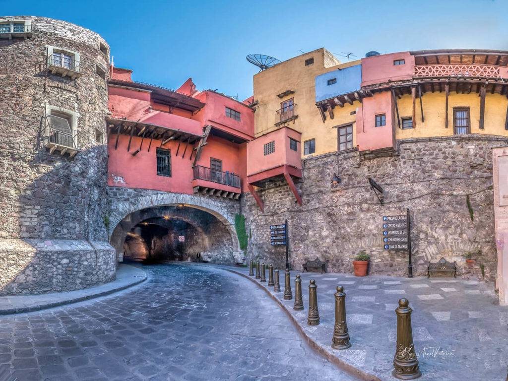 Rua de paralelepípedos em Guanajuato, México, com um túnel de pedra à esquerda e edifícios coloridos de tons rosa, amarelo e azul sobre uma base de pedra. Placas de rua indicam direções para pontos turísticos