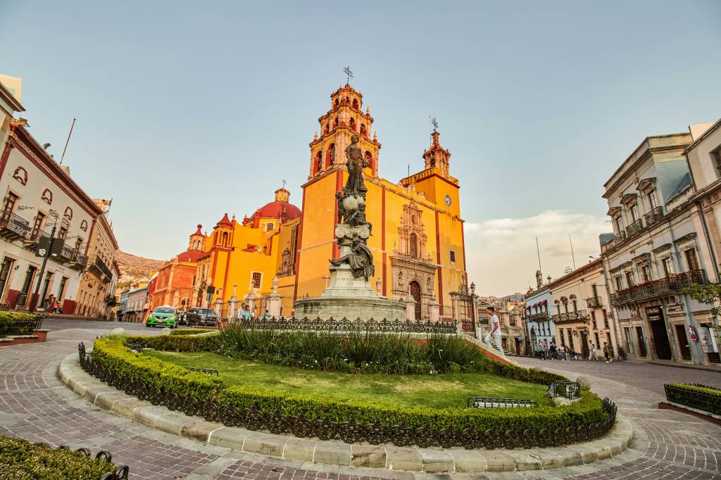 Praça com canteiro central de grama e arbustos, ladeada por ruas de paralelepípedos. Ao fundo, a Basílica Colegiada de Nossa Senhora de Guanajuato, de cor amarela e cúpulas vermelhas, com uma estátua escura em primeiro plano. Edifícios coloridos e carros estacionados completam a cena urbana sob um céu claro