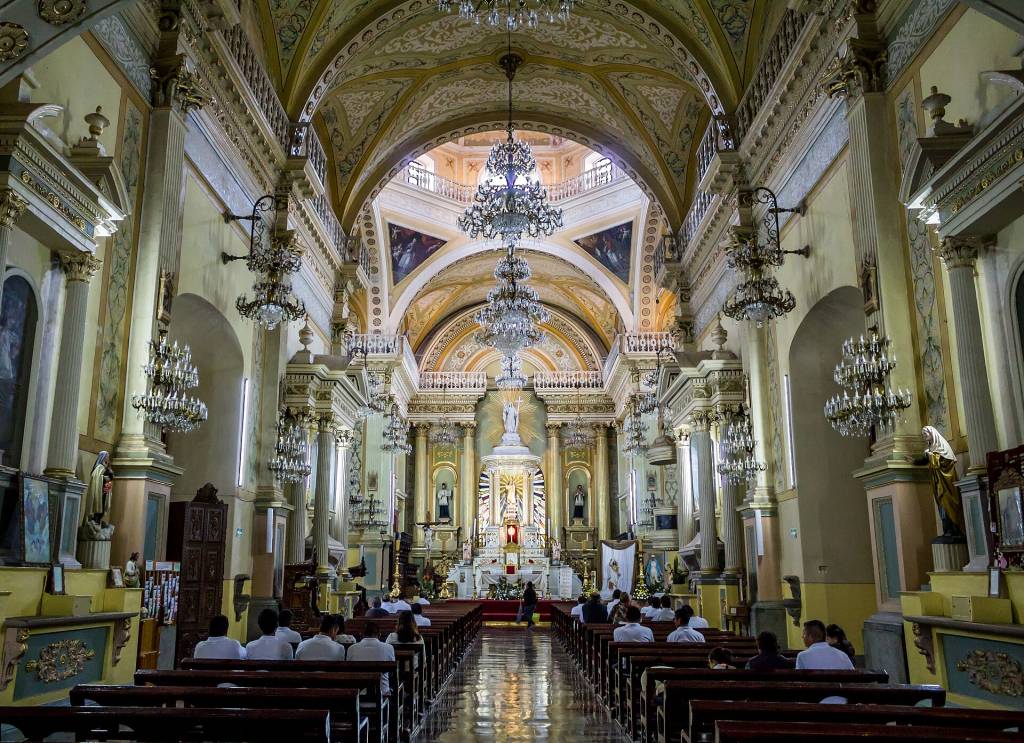 Interior de uma igreja católica ornamentada, com teto abobadado em tons de dourado e branco, lustres de cristal pendurados e colunas decoradas. Pessoas sentadas em bancos de madeira, de costas para a câmera, observam o altar principal ao fundo, iluminado e com uma estátua branca no centro. O chão reflete a luz, criando um caminho brilhante até o altar