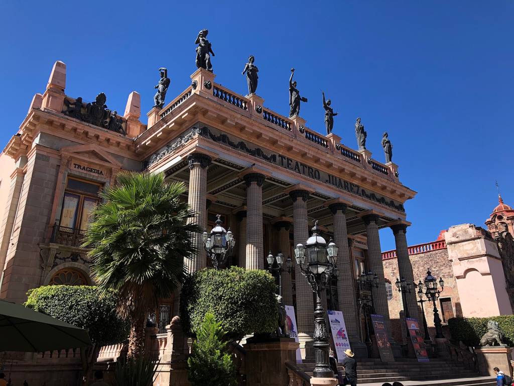 Fachada do Teatro Juárez em Guanajuato, México, com colunas coríntias, estátuas no telhado e letreiro TEATRO JUAREZ. O edifício cor-de-rosa tem a palavra TRAGEDIA acima de uma janela lateral, e há palmeiras e postes de luz ornamentados em primeiro plano, sob um céu azul claro