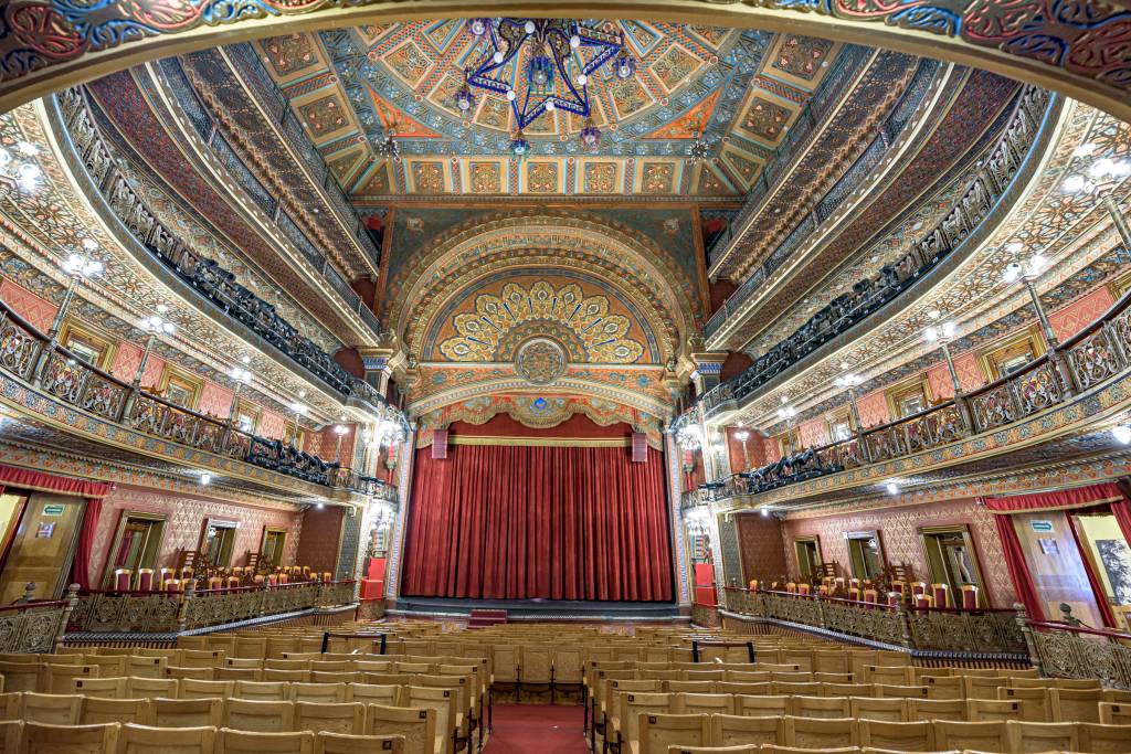 Interior de um teatro antigo com poltronas de madeira vazias no térreo e balcões ornamentados em vários níveis. O palco tem uma cortina vermelha fechada, e o teto e paredes são ricamente decorados com padrões geométricos e florais em tons de dourado, azul, laranja e vermelho. Lustres pendem do teto, iluminando o ambiente luxuoso