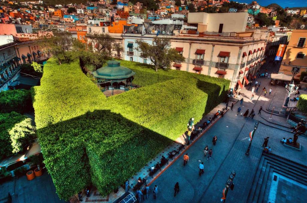 Vista aérea de um parque com arbustos altos e densos formando um labirinto verde, com um coreto verde-escuro no centro. Pessoas caminham pelas ruas de paralelepípedos ao redor, ladeadas por edifícios coloridos e casas em uma colina ao fundo, sob luz solar intensa