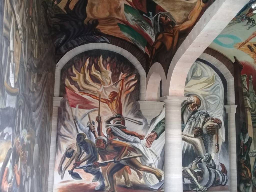 Mural com arcos brancos, retratando cenas de luta e sofrimento. À esquerda, figuras com ferramentas e bandeiras vermelhas, sob chamas. À direita, uma mulher de branco segura um corpo, enquanto outra figura jaz acorrentada no chão. O teto exibe mais figuras em tons terrosos e escuros