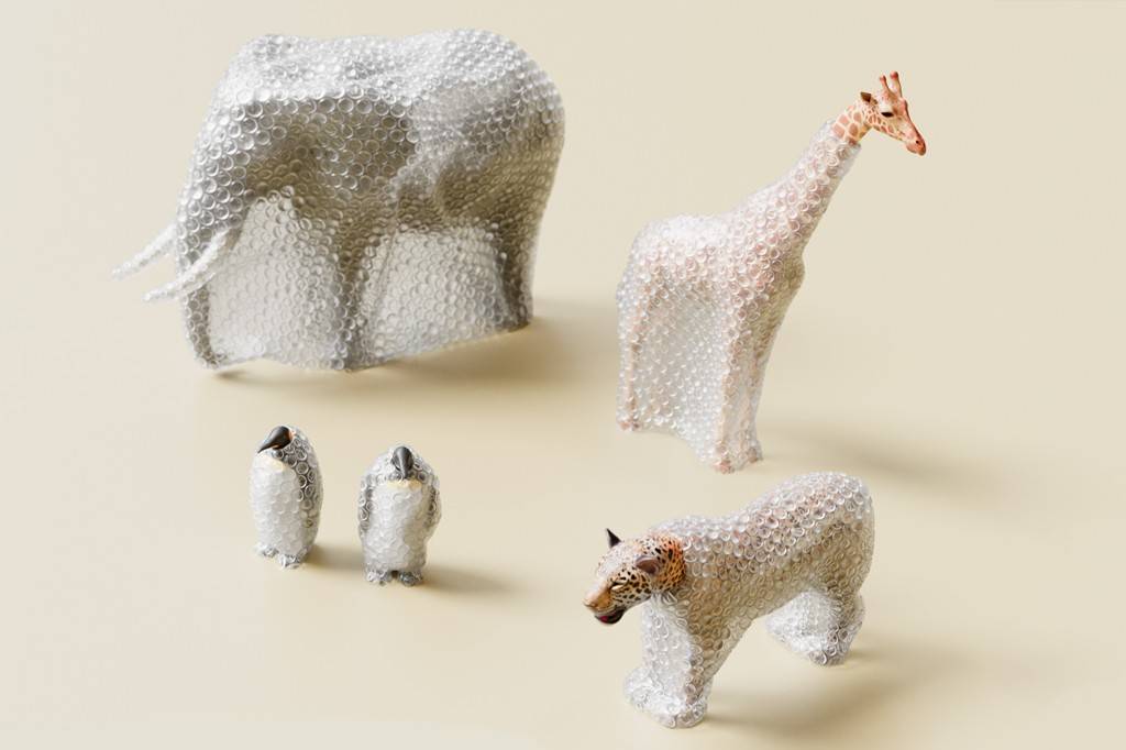 SI_488_Zoológico_1 Estatuetas de animais embrulhadas em plástico bolha: um elefante, uma girafa, dois pinguins e um leopardo, com as cabeças e detalhes expostos, sobre um fundo bege claro.
