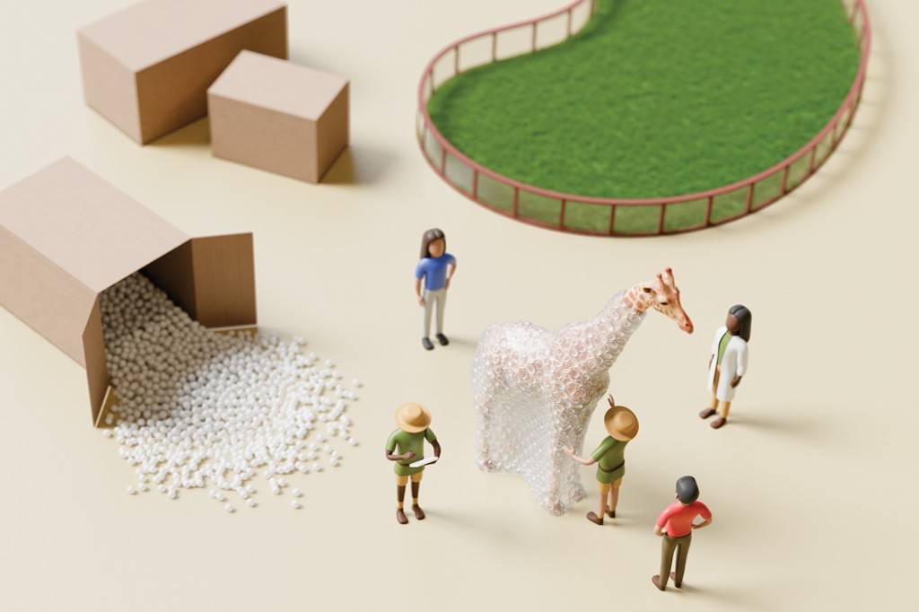 SI_488_Zoológico_3 Miniaturas de pessoas e objetos em um cenário bege. Uma girafa coberta por plástico bolha está no centro, com um homem de chapéu e roupa verde tocando-a. Outro homem de chapéu e roupa verde segura um caderno. Uma mulher de camiseta azul e calça clara, uma mulher de jaleco branco e um homem de camisa vermelha e calça marrom observam. À esquerda, uma caixa de papelão derrama bolinhas brancas, e outras duas caixas fechadas estão próximas. No canto superior direito, um cercado verde com grama simula um habitat.
