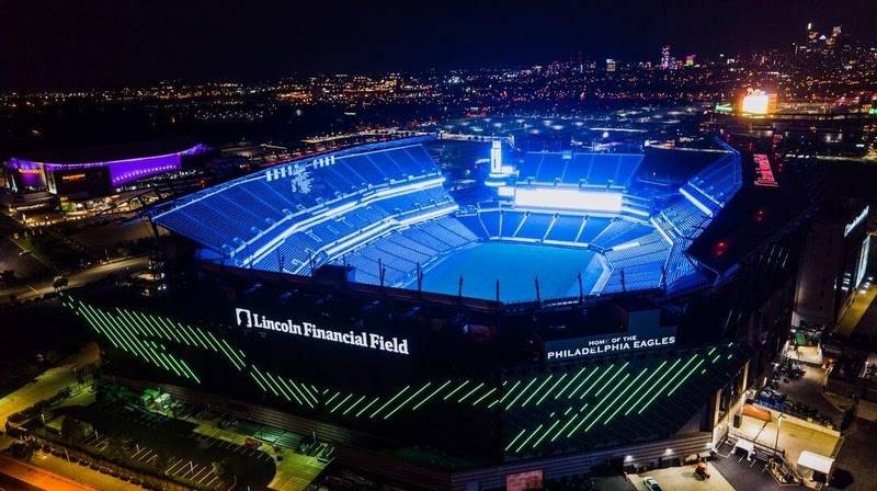 Vista aérea noturna do Lincoln Financial Field, estádio do Philadelphia Eagles, iluminado em azul vibrante. O campo e as arquibancadas brilham em azul, enquanto a fachada exibe o nome do estádio e HOME OF THE PHILADELPHIA EAGLES em branco, com linhas verdes diagonais. Ao fundo, a cidade se estende com luzes cintilantes
