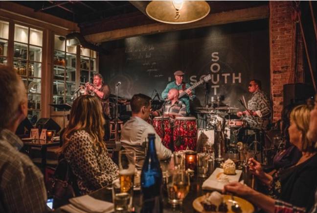 Pessoas assistem a uma banda de jazz tocando em um bar com iluminação baixa. Uma mulher toca trompete, um homem toca baixo, outro congas e um quarto bateria. Clientes sentados em mesas, com bebidas e pratos, observam a apresentação. O ambiente é acolhedor, com paredes de tijolos e prateleiras de madeira.