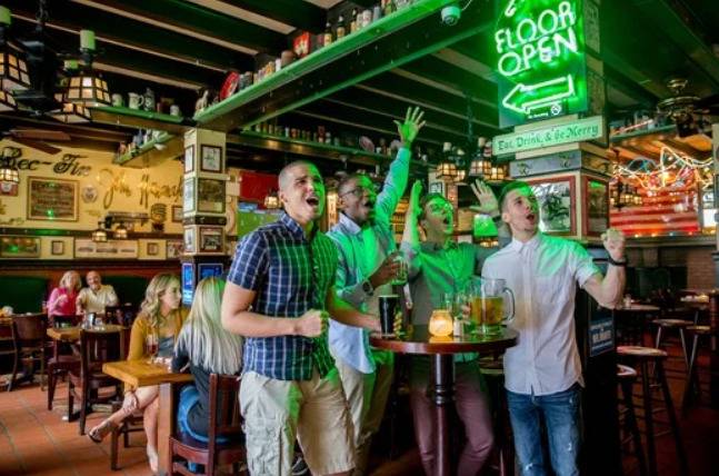 Grupo de homens em um bar, comemorando e gritando, alguns com os braços levantados. Um letreiro neon verde FLOOR OPEN ilumina o ambiente, que tem mesas de madeira e prateleiras com garrafas