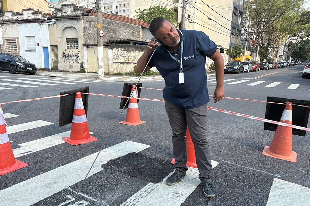 Homem de pele parda, barba e camiseta azul da Sabesp, inclina-se para ouvir um aparelho auditivo enquanto segura uma bengala longa. Ele está em uma rua com faixa de pedestres, cones de sinalização e fita zebrada, indicando obras. Ao fundo, carros estacionados e prédios antigos.