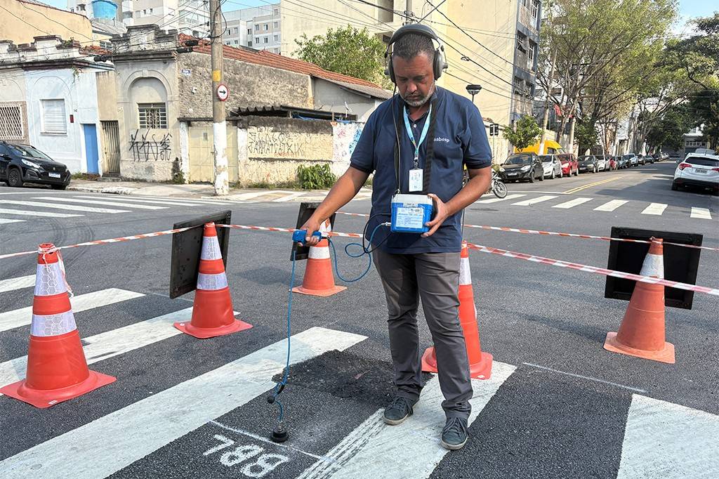 Homem de barba, camisa polo azul e fones de ouvido, usa equipamento para detectar vazamentos em rua com faixa de pedestres e cones de sinalização