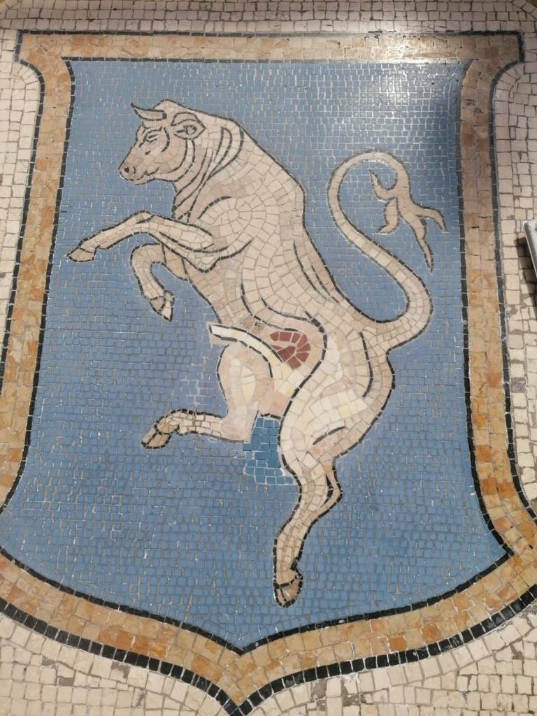 Mosaico de um touro branco empinado sobre um escudo azul claro, com borda marrom e preta. O touro tem chifres, cauda enrolada e um órgão genital vermelho visível