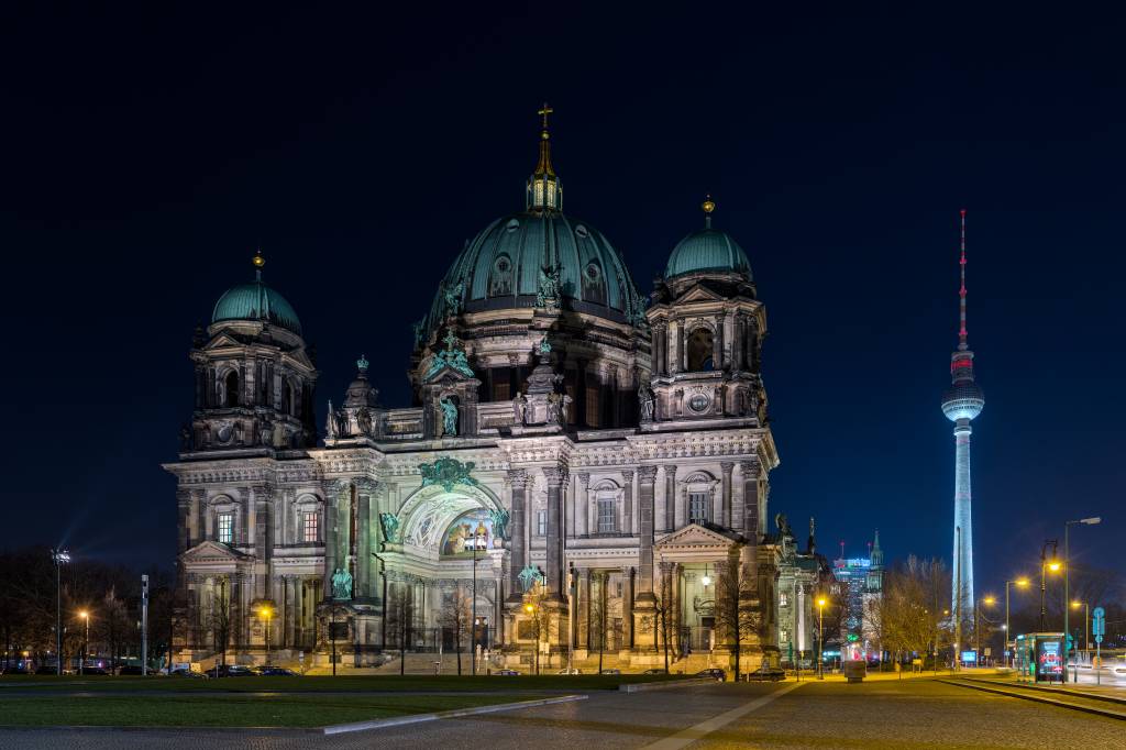 Catedral de Berlim iluminada à noite, com cúpulas verdes e detalhes arquitetônicos em pedra clara. À direita, a Torre de TV de Berlim brilha com luzes azuis e vermelhas, destacando-se no céu escuro. Ruas e árvores com iluminação amarela no primeiro plano