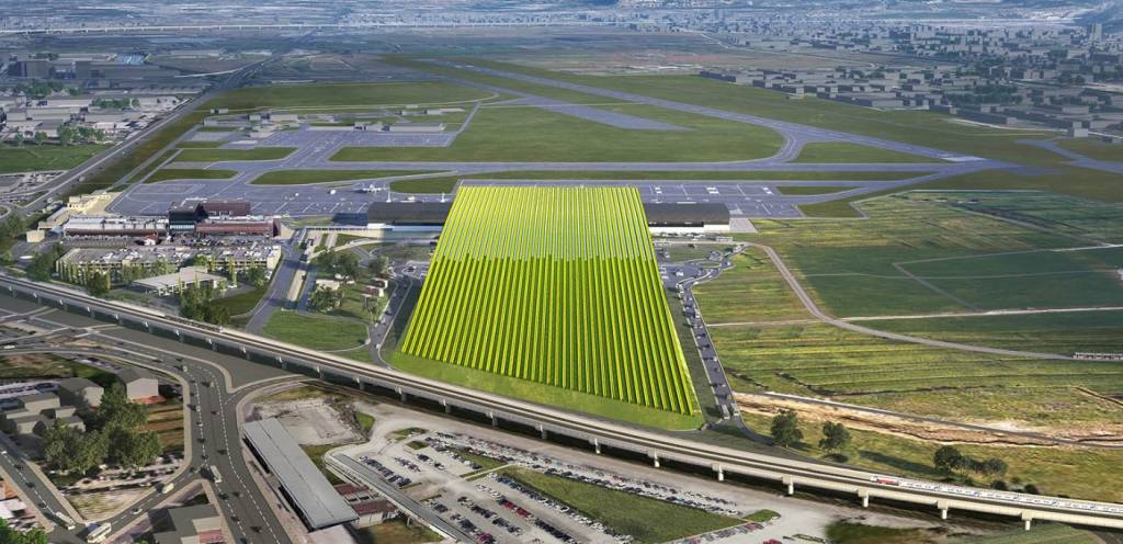 Vista aérea de um aeroporto com pistas de pouso e decolagem, edifícios de terminais e um grande campo verde com painéis solares. Há uma rodovia e uma linha de trem elevadas, com carros e um trem visíveis. Ao fundo, uma área urbana com edifícios.