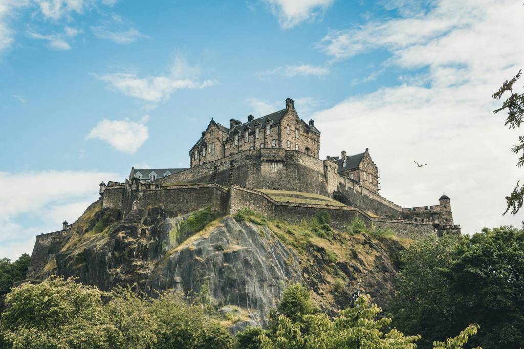 Castelo de Edimburgo, Escócia, construído em pedra cinza, com telhados escuros e várias janelas, erguido sobre uma colina rochosa coberta por vegetação verde. O céu azul claro tem nuvens esparsas e um pássaro voa à direita. Árvores frondosas aparecem em primeiro plano