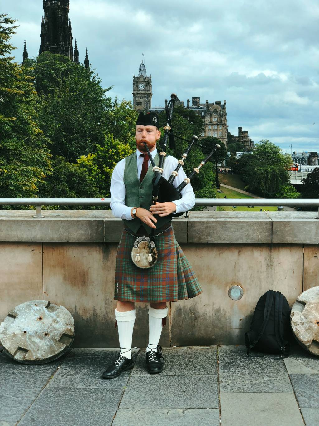 Homem ruivo com barba, vestindo kilt xadrez verde e marrom, colete verde e camisa branca, tocando gaita de fole em Edimburgo, Escócia. Ao fundo, árvores e edifícios históricos sob céu nublado