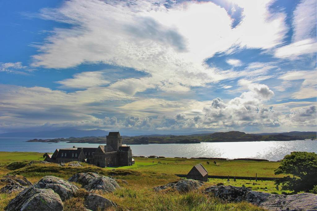 Antiga abadia de pedra cinza com torre, em uma ilha verdejante, com um lago azul-prateado e montanhas ao fundo, sob céu azul com nuvens brancas e cinzas