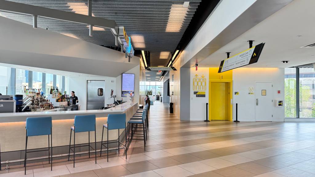 Interior moderno de um bar com balcão branco e bancos azuis, onde uma barista atende. Ao lado, um corredor com piso listrado e elevadores amarelos, além de sinalização de direção para Baggage Claim e Valet Parking. Grandes janelas mostram prédios e árvores ao fundo