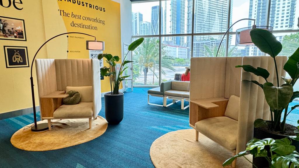 Ambiente de coworking moderno com paredes amarelas e carpete azul. Duas poltronas individuais bege com laterais altas e mesa de madeira, sobre tapetes ovais amarelos. Plantas verdes em vasos pretos e luminárias de chão arqueadas complementam o espaço. Ao fundo, uma grande janela revela prédios altos e palmeiras, com uma pessoa sentada em um sofá cinza